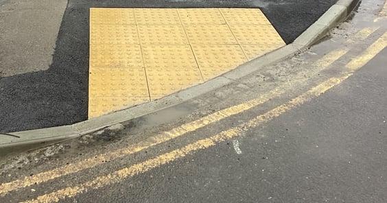 A close up picture of a dropped kerb in the UK, the tarmac has been re-done recently and tactile paving leads up to the edge of the kerb