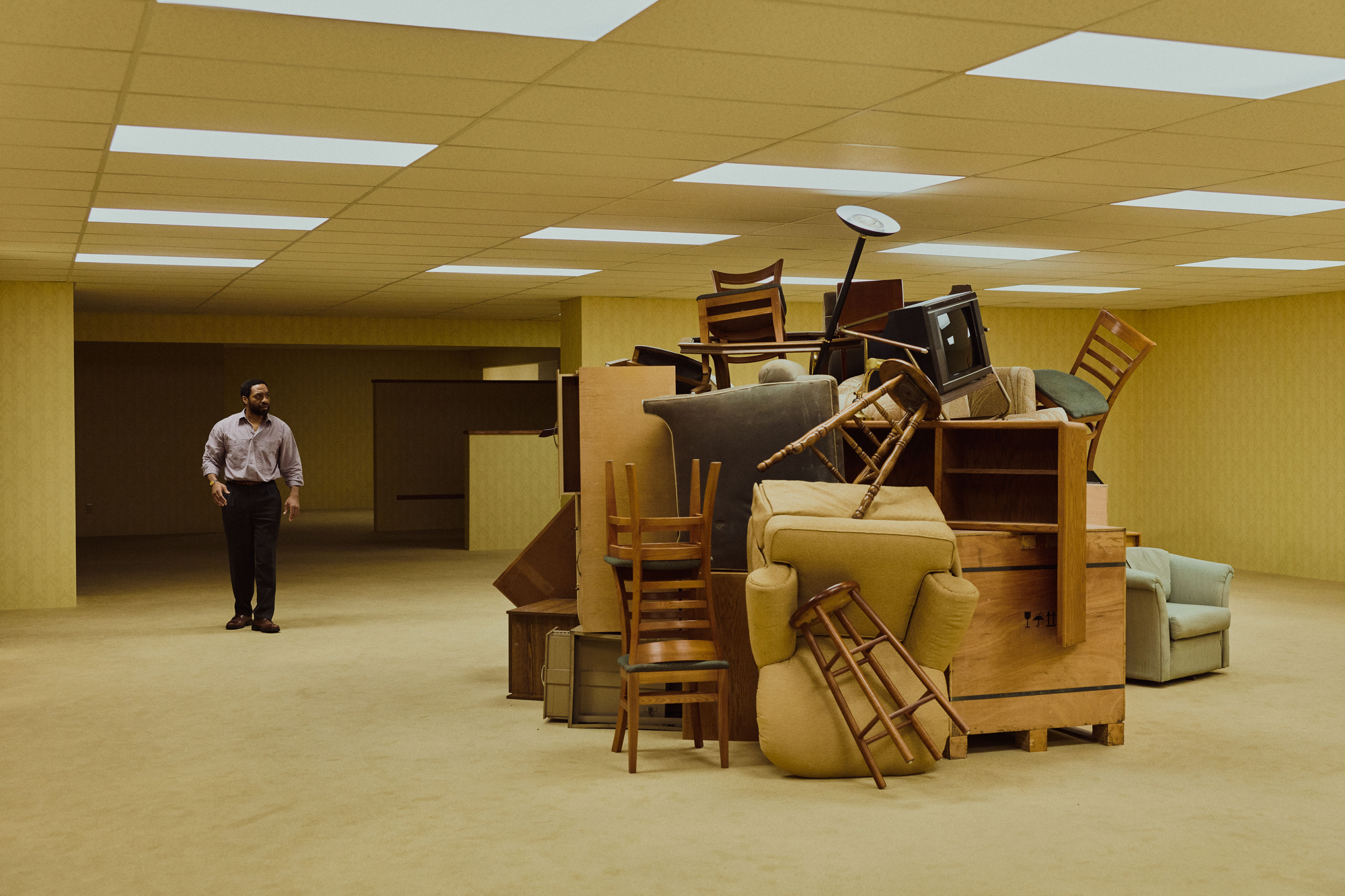 A still from the film The Backrooms: a man stands alone in an unnervingly empty fluorescent office space, facing a chaotic, towering pile of furniture — the kind of ordinary place made strange by having no clear purpose or exit.